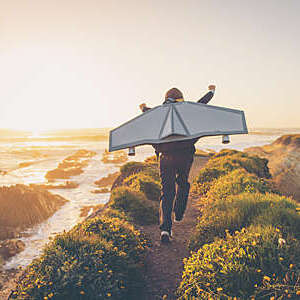 A young boy dressed in business suit and tie wears a homemade jetpack and flying goggles raises his arms in the afternoon sun while running to take off into the air on an outcropping above the surf in California. This young entrepreneur is ready to take his new business to new heights.