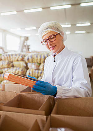 Smiling female worker in sterile lab coat packs finished food products in boxes in a food factory.
