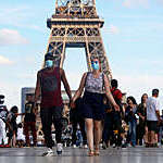 Tourist couple wearing protective masks walk along the Eiffel Tower esplanade in Paris holding hands.