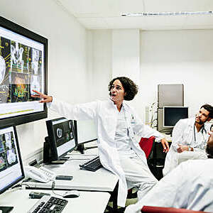 A clinical registrar reviewing a patients test results, looking at some information on a large monitor with some other doctors.